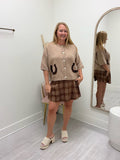 A woman modeling a beige knit cropped cardigan with black horseshoe details and a brown plaid mini skirt, standing in a fitting room with a mirror.