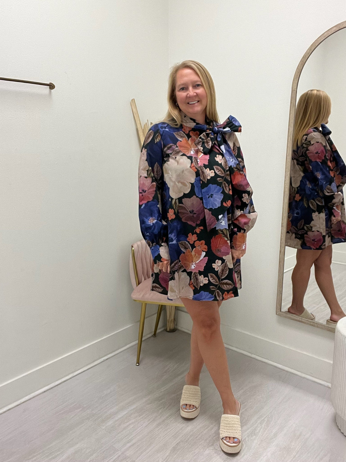 A woman models a long-sleeve floral mini dress with a large bow at the collar in a boutique setting. The dress features a multicolor floral pattern on a dark navy background.