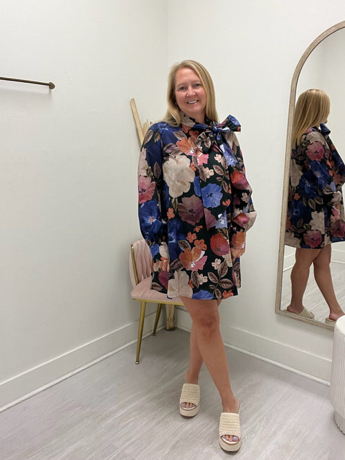 A woman models a long-sleeve floral mini dress with a large bow at the collar in a boutique setting. The dress features a multicolor floral pattern on a dark navy background.