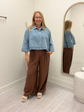 A woman stands in a fitting room wearing a cropped light blue denim jacket and high-waisted brown wide-leg pants, with her reflection visible in an arched mirror.