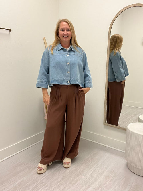 A woman stands in a fitting room wearing a cropped light blue denim jacket and high-waisted brown wide-leg pants, with her reflection visible in an arched mirror.