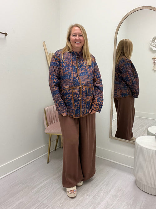 A woman stands in a boutique wearing a blue and orange patterned long-sleeve crop top with a tie waist, paired with wide-leg brown pants. She is standing in front of a large arched mirror that reflects her back, with a pink chair and light wood flooring visible in the room.