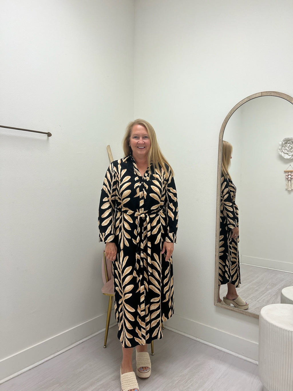A woman stands in a fitting room wearing a long-sleeve, button-front maxi dress with a black and beige leaf pattern. She is standing next to a large arched mirror that reflects her, with a light gray floor and white walls.
