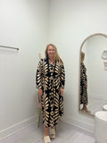 A woman stands in a fitting room wearing a long-sleeve, button-front maxi dress with a black and beige leaf pattern. She is standing next to a large arched mirror that reflects her, with a light gray floor and white walls.