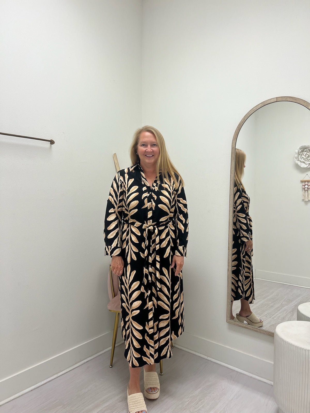 A woman stands in a fitting room wearing a long-sleeve, button-front maxi dress with a black and beige leaf pattern. She is standing next to a large arched mirror that reflects her, with a light gray floor and white walls.