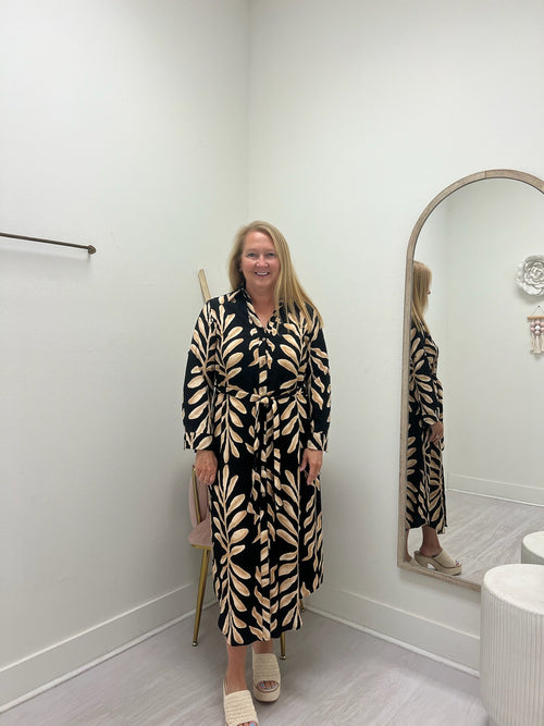 A woman stands in a fitting room wearing a long-sleeve, button-front maxi dress with a black and beige leaf pattern. She is standing next to a large arched mirror that reflects her, with a light gray floor and white walls.